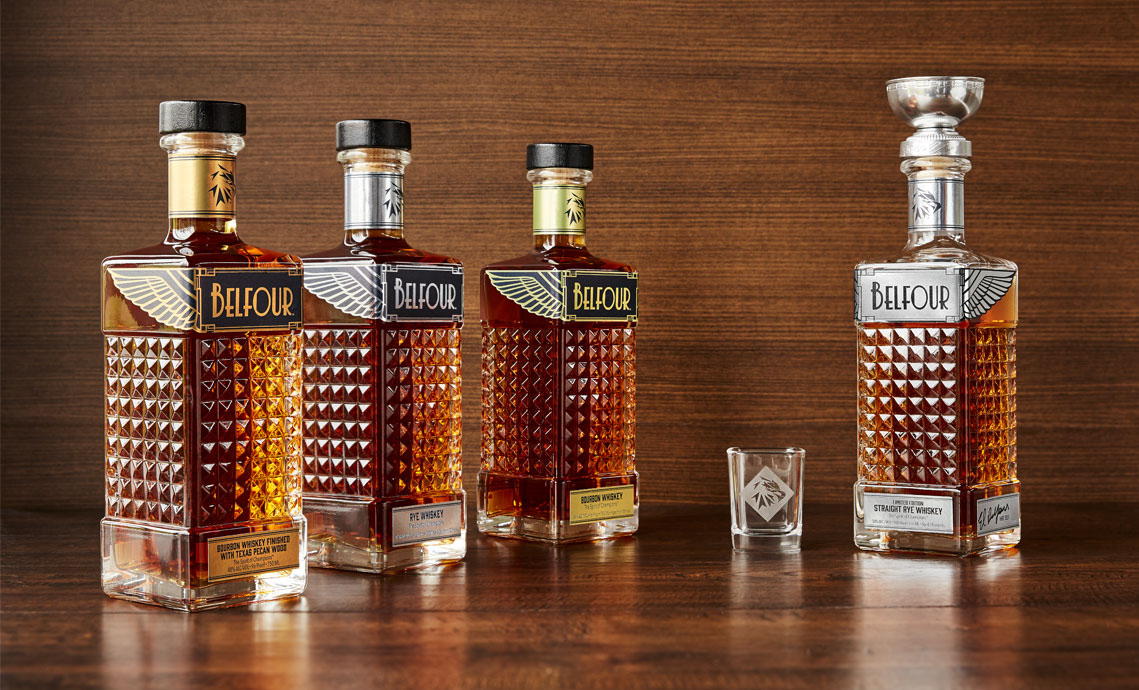 Group shot of Texas Pecan Whiskey, Rye Whiskey, Bourbon and Limited Edition Rye whiskey on polished wood with a wood backdrop and a Belfour-branded shot glass